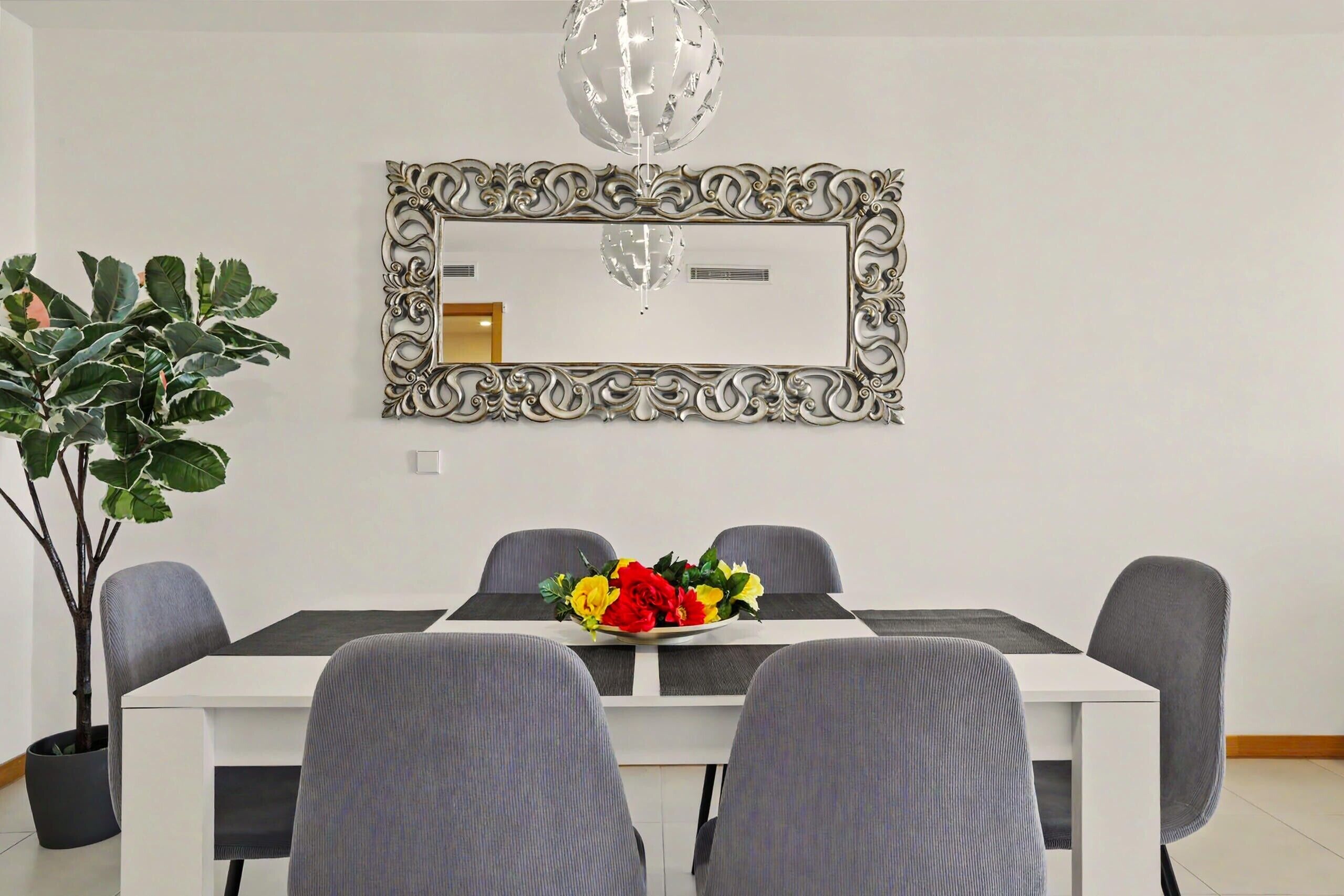 A dining area with a white table, grey chairs, a decorative mirror, and a potted plant.