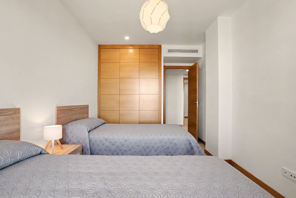 A bedroom with two beds, a wooden wardrobe, a nightstand with a lamp, and a ceiling light fixture.