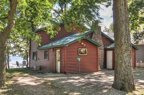 A rustic cabin with a stone chimney, a wooden door, and a sign reading 'The Esser Den.'