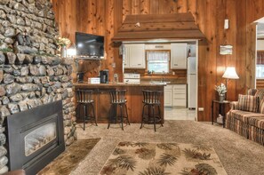 Un interior acogedor con pared de piedra, chimenea, barra con taburetes, cocina al fondo y sofá con cojines a cuadros.
