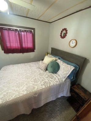 A bedroom with a bed, a wall clock, a window with curtains, and a small wooden nightstand.