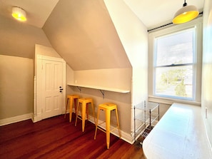 A room with a sloped ceiling, a white door, wooden stools, a white shelf, a window with blinds, and a ceiling light. - Caldwell's Attic — Cozy King Bed Attic Getaway + Central Location (Staunton)