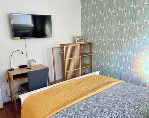 Un dormitorio con cama, televisor montado en la pared, escritorio con lámpara y estantería.