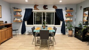 Un coin repas avec une table et des chaises, une fenêtre avec des rideaux, une bibliothèque et un meuble orné de plantes.