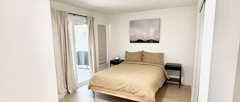 A bedroom with a bed, a nightstand, a wall-mounted lamp, and a window with curtains.