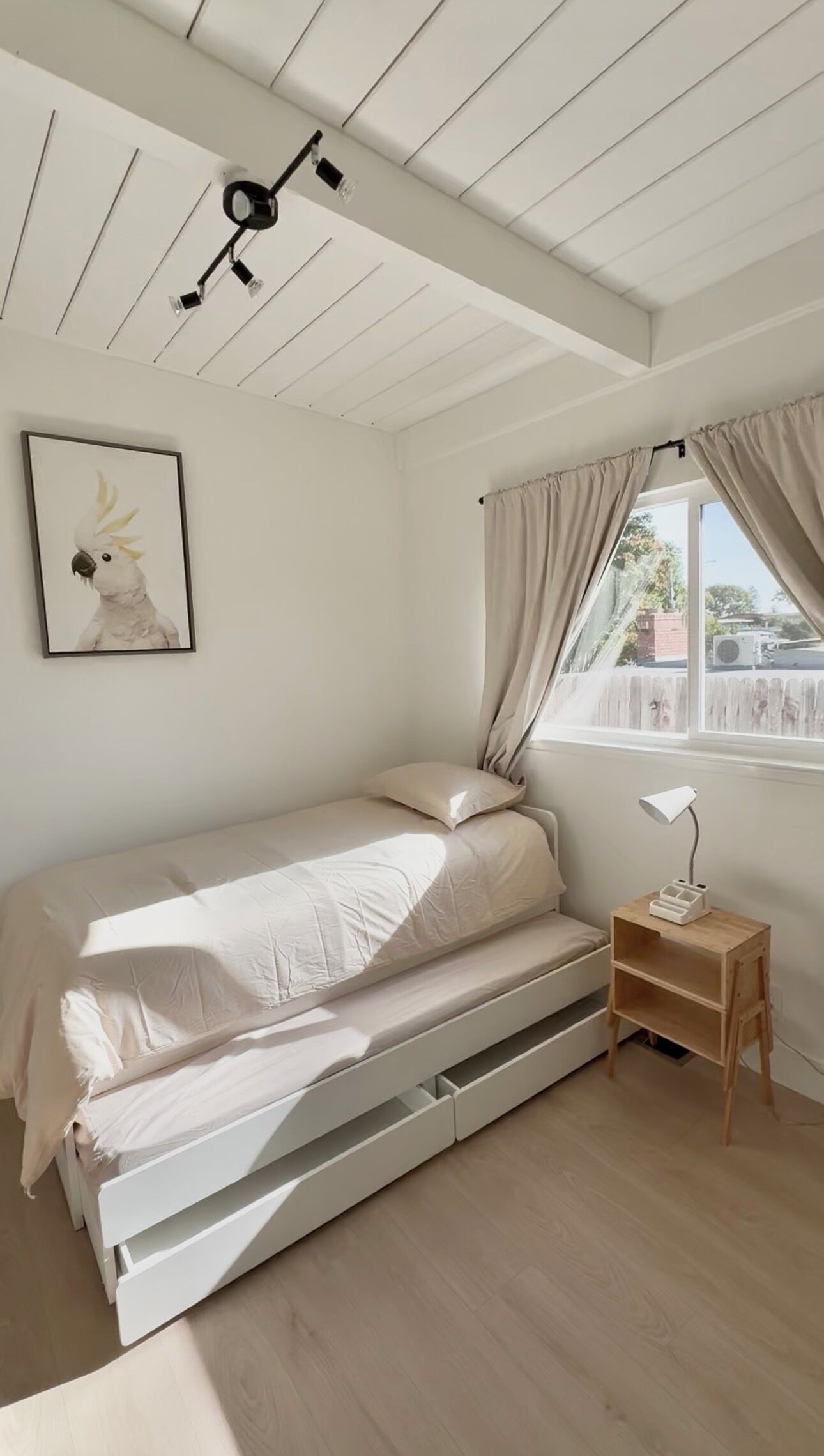 Quarto minimalista com cama, abajur, uma pequena mesa de cabeceira de madeira e uma janela com cortinas.