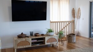 Un soggiorno con una televisione a schermo piatto montata a parete, un tavolino consolle in legno con oggetti decorativi, una scala con ringhiera in legno e un'ampia finestra con tende leggere.