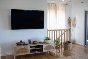 A living room with a flat-screen TV mounted on the wall, a wooden console table with decorative items, a staircase with a wooden railing, and a large window with sheer curtains. - Quampi – Unbelievable Main Beach Views (Point Lookout)