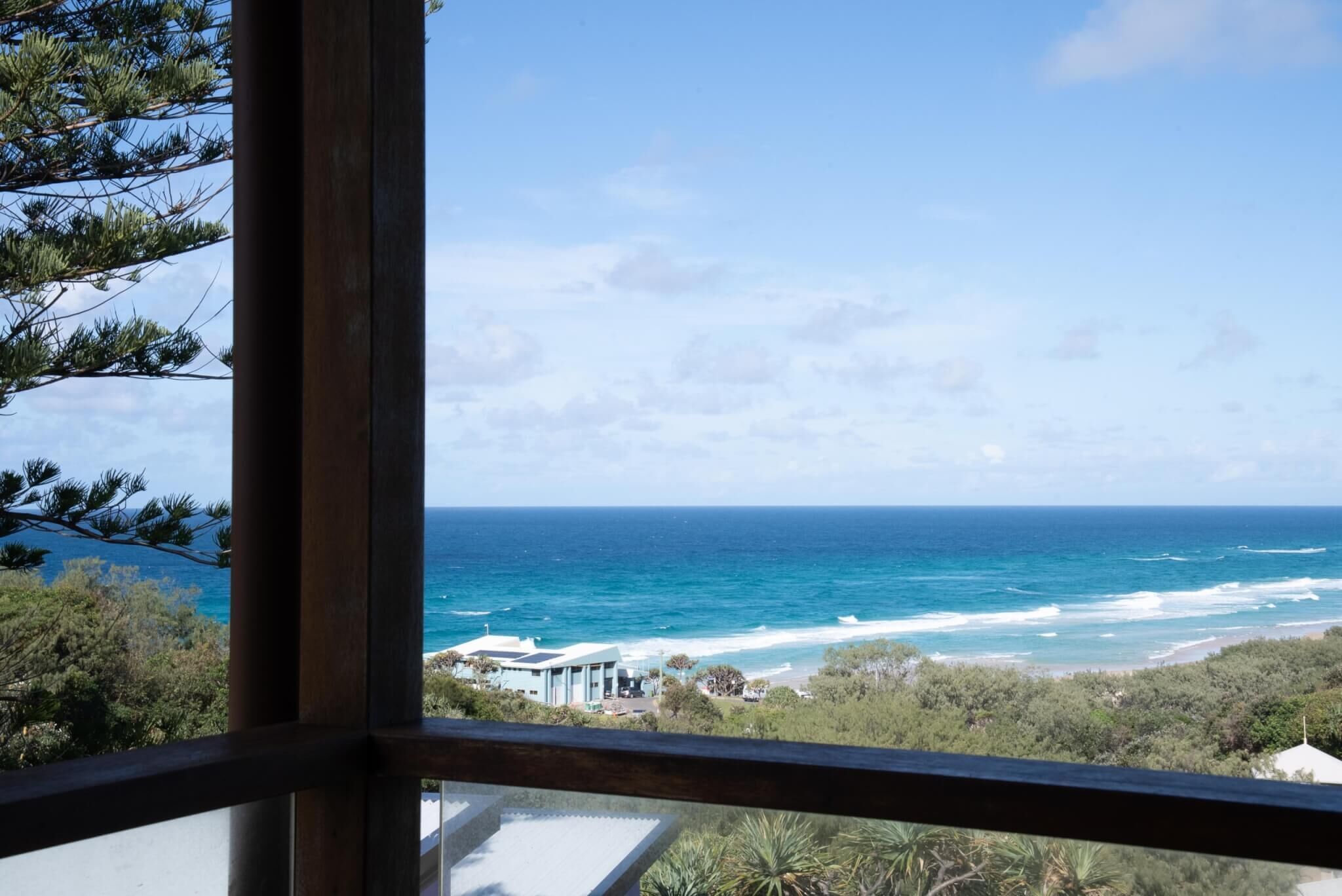 A coastal view through a window, featuring a beach, ocean, and a building with a modern design.