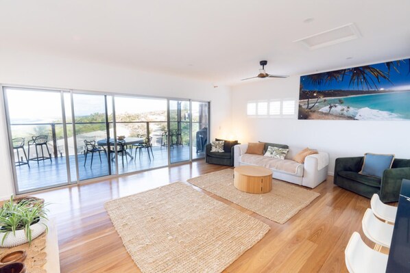 A spacious living area with a large sliding glass door leading to an outdoor deck, a ceiling fan, and a painting of a beach scene. - Quampi – Unbelievable Main Beach Views (Point Lookout)