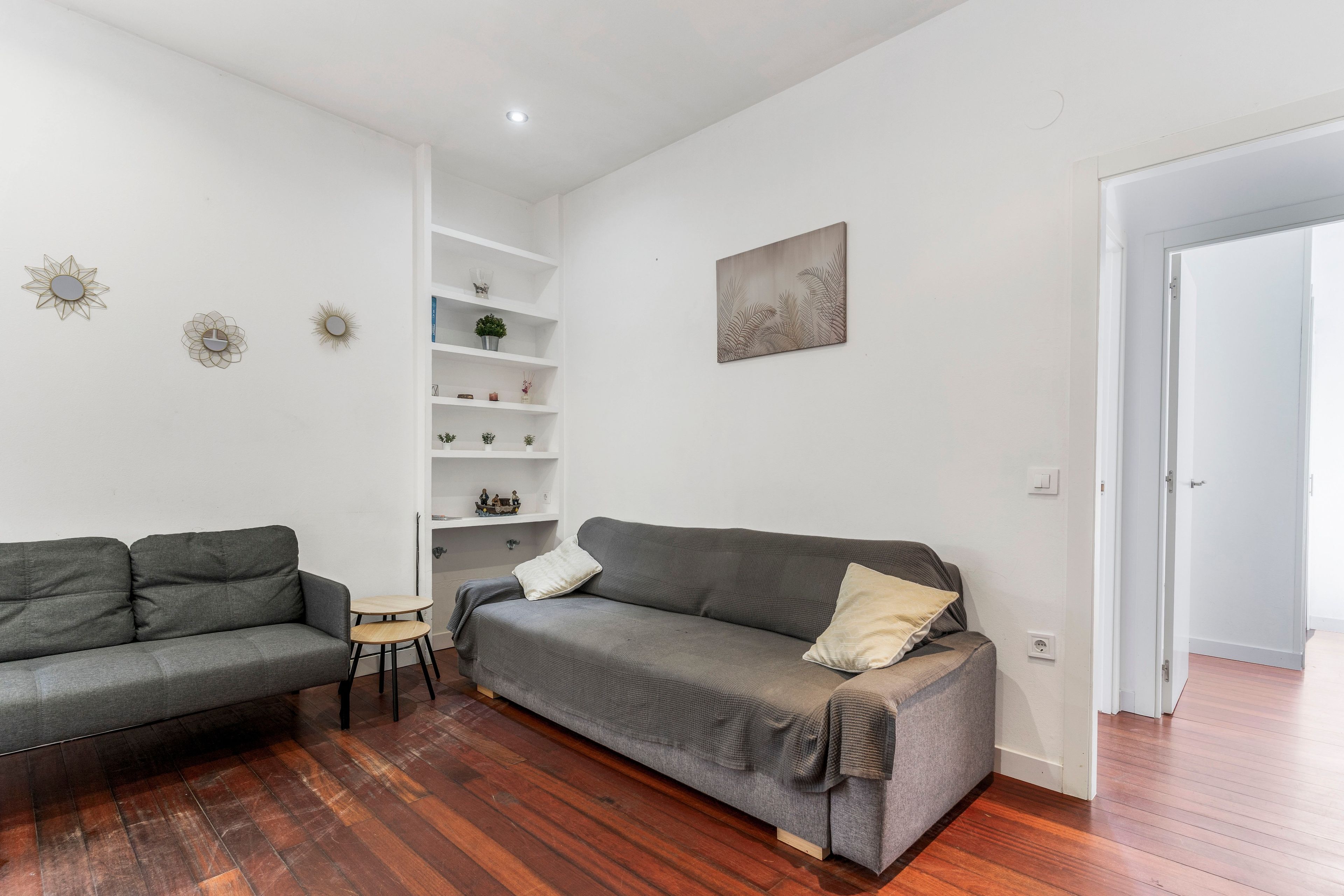 Un salon moderne avec un canapé gris, un plus petit canapé gris, une table basse en bois et une étagère blanche agrémentée d’objets décoratifs.