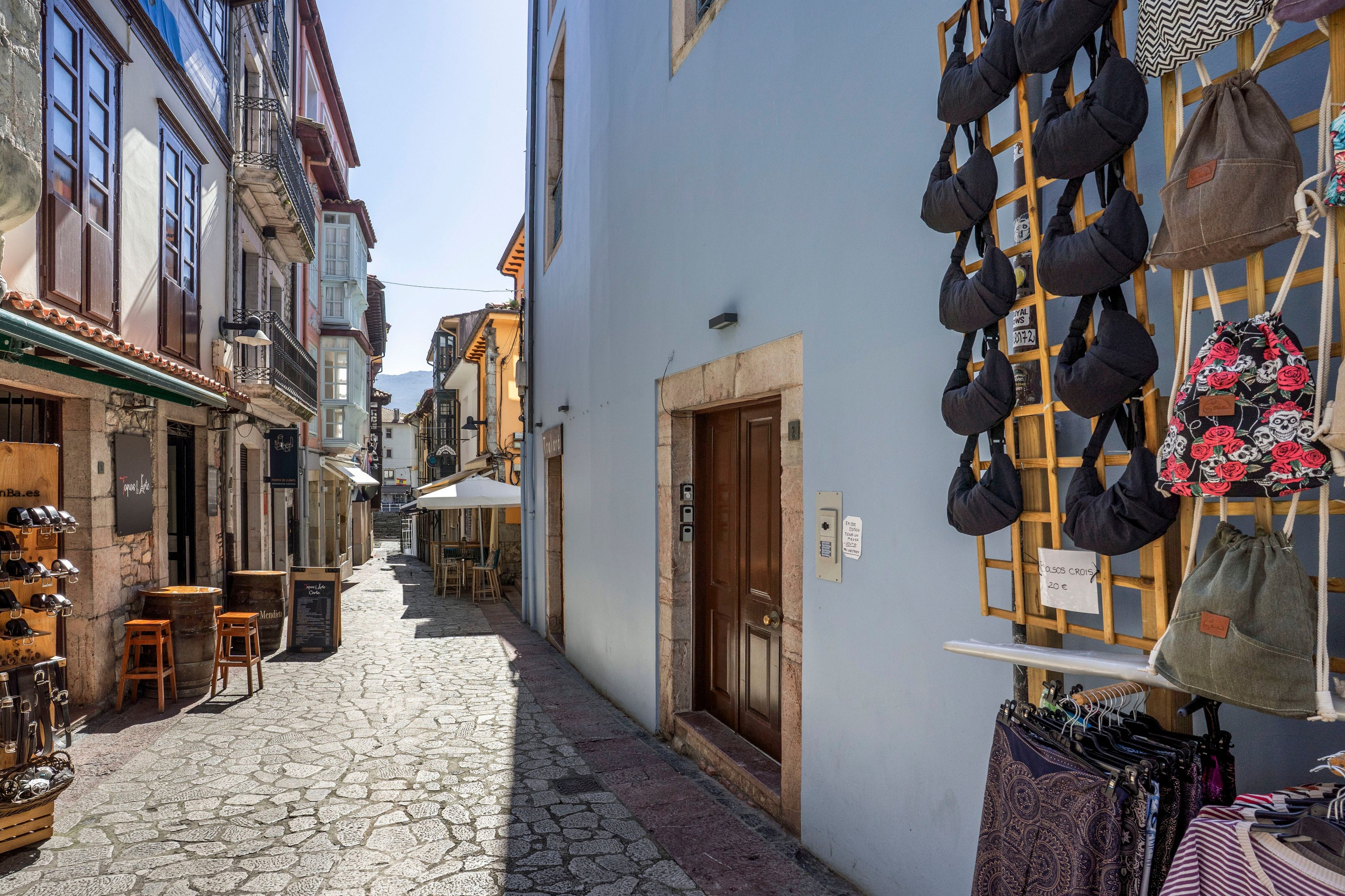 Une ruelle étroite pavée de pierres, bordée de bâtiments des deux côtés, avec une porte en bois et un présentoir à sacs.