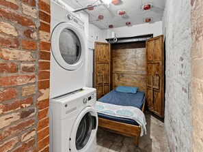 A small bedroom with a washing machine, a bed with a blue pillow, and wooden wardrobes.