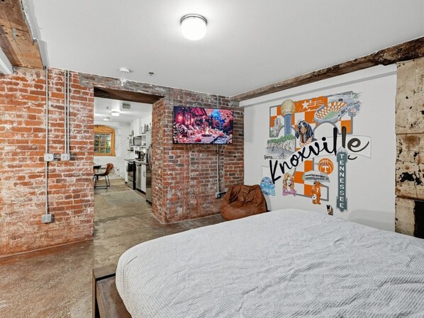 A room with a bed, a brick wall, a TV mounted on the wall, and a wall with a Knoxville logo.