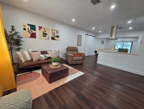 A living room with a brown leather sofa, a brown armchair, a coffee table with a plant, and a kitchen area in the background.