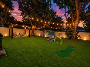 A backyard with artificial lighting, a green lawn, a golf putting area, and a covered seating area.