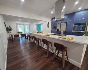 A modern kitchen with a central island, bar stools, a dining area with a table and chairs, and a view of the outdoors.