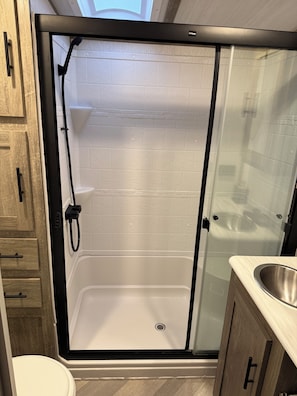 Un box de ducha con puerta de cristal, pared revestida de azulejos blancos y mueble de madera con lavabo.