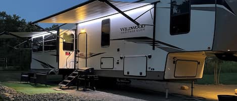 Una caravana Wildwood Heritage Glen estacionada en un camino de grava con un área exterior cubierta y un pequeño porche.