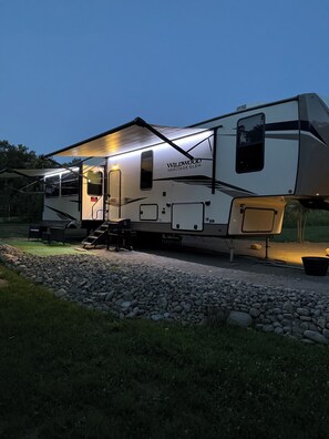 Una caravana Wildwood Heritage Glen estacionada en un camino de grava con un área exterior cubierta y un pequeño porche.