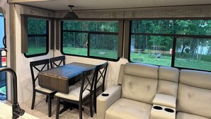 A compact RV interior with a dining area, cushioned seating, and large windows offering a view of a wooded area.