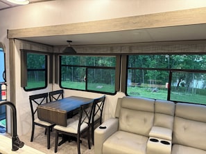 A compact RV interior with a dining area, cushioned seating, and large windows offering a view of a wooded area. - Fabulous 3-bedroom RV with direct access to the Sebasticook River! (Benton)