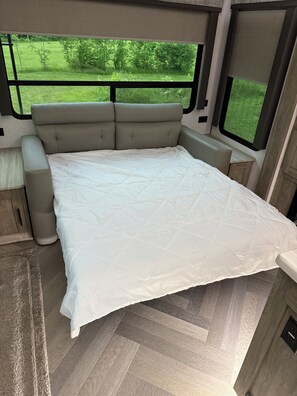 A bed with a white quilt in an RV with a view of greenery outside.