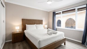 Un dormitorio ordenado con cama, lámparas de noche, mesitas de noche, ventilador de techo y ventana con cortinas.