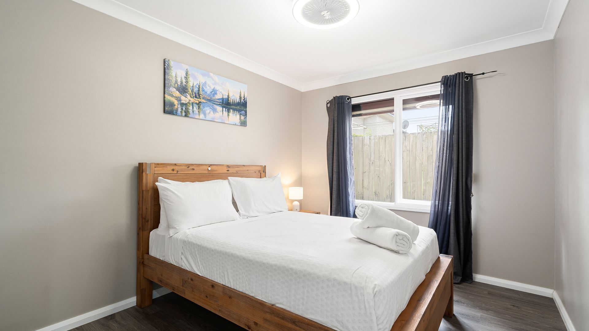 A bedroom with a wooden bed, white bedding, a nightstand with a lamp, a painting on the wall, and a window with curtains.