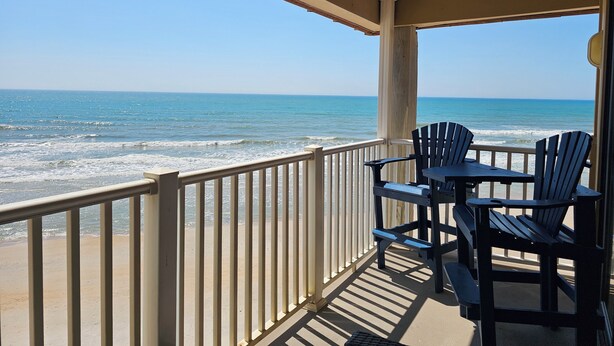 A covered outdoor area with a view of the ocean, a railing, and two Adirondack chairs.