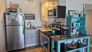 A compact kitchen with a stainless steel refrigerator, a microwave, a coffee maker, and a teal-colored shelf with mugs and jars.