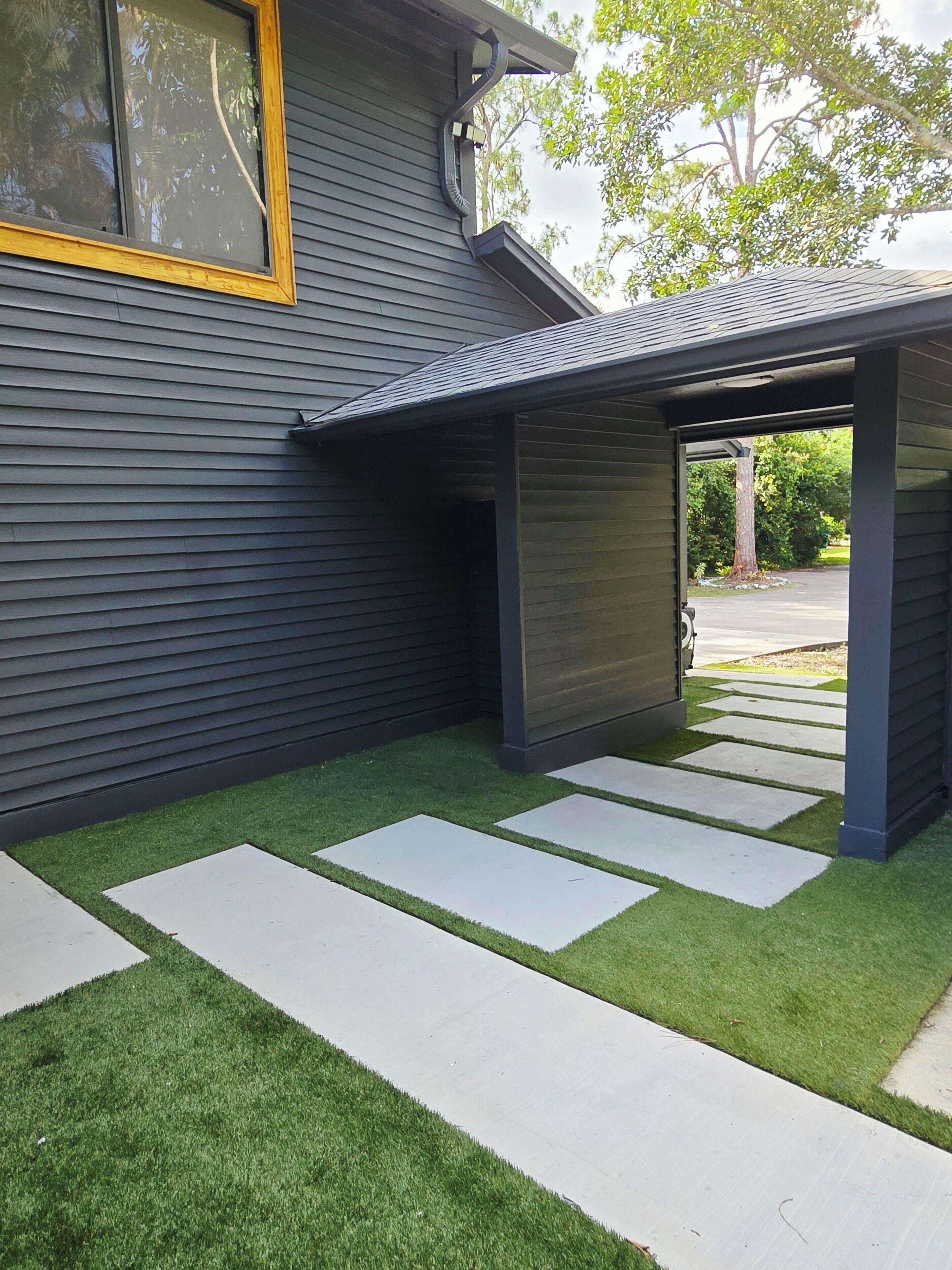 A modern house with a dark exterior, a window with a yellow frame, and a covered patio area with artificial grass and stone pathways.