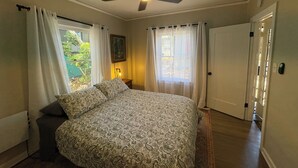 A bedroom with a bed, a ceiling fan, a window with curtains, a door, and a nightstand with a lamp.