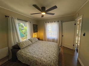 Une chambre à coucher avec un lit, un ventilateur de plafond, une fenêtre avec des rideaux, une porte et une table de chevet avec une lampe.