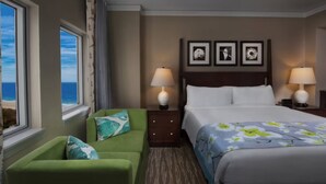 Ein Hotelzimmer mit einem Bett, zwei Nachttischlampen, einem grünen Sessel, einem Fenster mit Meerblick und drei gerahmten Bildern an der Wand.