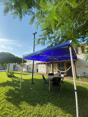 A covered outdoor seating area with chairs and a table, surrounded by a well-maintained lawn and a building with windows. - Your home away from home (Acapulco de Juárez)