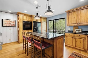 A kitchen with wooden cabinets, a black refrigerator, a large island with bar stools, pendant lights, and a window with a view of trees. - Warm Springs Ranch Hideaway | Pool | Ski Access | Mtn. Views (Ketchum)
