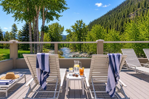 A deck with lounge chairs, a table with drinks, and a view of a river and forest. - Warm Springs Ranch Hideaway | Pool | Ski Access | Mtn. Views (Ketchum)