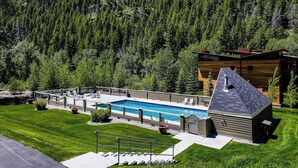 En modern byggnad med en pool, omgiven av en välskött gräsmatta och en skogbeklädd bergssluttning i bakgrunden.