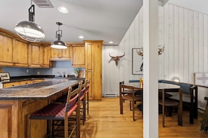 A kitchen with wooden cabinets, a central island with stools, and a dining table with chairs. - Warm Springs Ranch Hideaway | Pool | Ski Access | Mtn. Views (Ketchum)