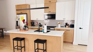 Cocina moderna con una isla central, armarios de madera y electrodomésticos de acero inoxidable, junto con dos taburetes negros.