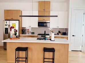 A modern kitchen with wooden cabinets, a stainless steel refrigerator, a microwave, and a gas stove. There are two black stools at the island.