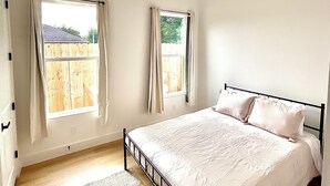 Un dormitorio con cama, ventilador de techo, dos ventanas con cortinas y vista a una cerca de madera.