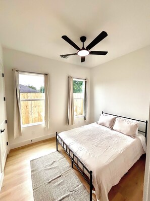 Ein Schlafzimmer mit einem Bett, einem Deckenventilator, zwei Fenstern mit Vorhängen und Blick auf einen Holzzaun.
