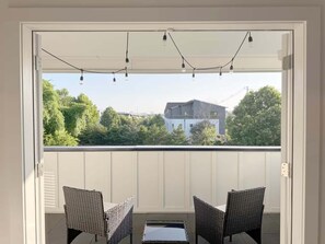 A balcony with two wicker chairs, a table, and a view of a house and trees. - Lookout Living 1405–Downtown Access–Sleeps 8 (Chattanooga)
