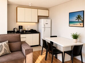 Une cuisine moderne avec des armoires blanches, un comptoir noir et un réfrigérateur. On y trouve une table à manger avec quatre chaises et une plante en pot. Un canapé avec un coussin à motifs se trouve au premier plan.
