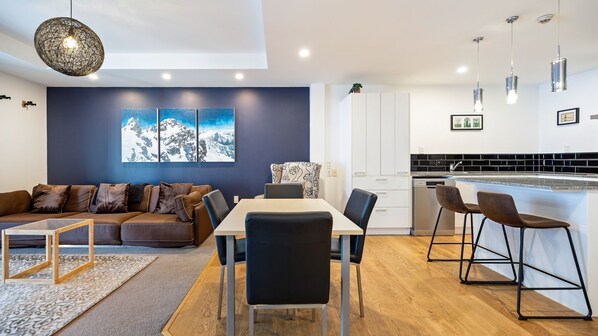 Ein modernes Wohnzimmer mit einer Couch, einem Esstisch und Stühlen, eine Küche mit weißen Schränken und einer schwarzen Spritzschutzwand, und ein großes Fenster mit Blick auf die Berge.