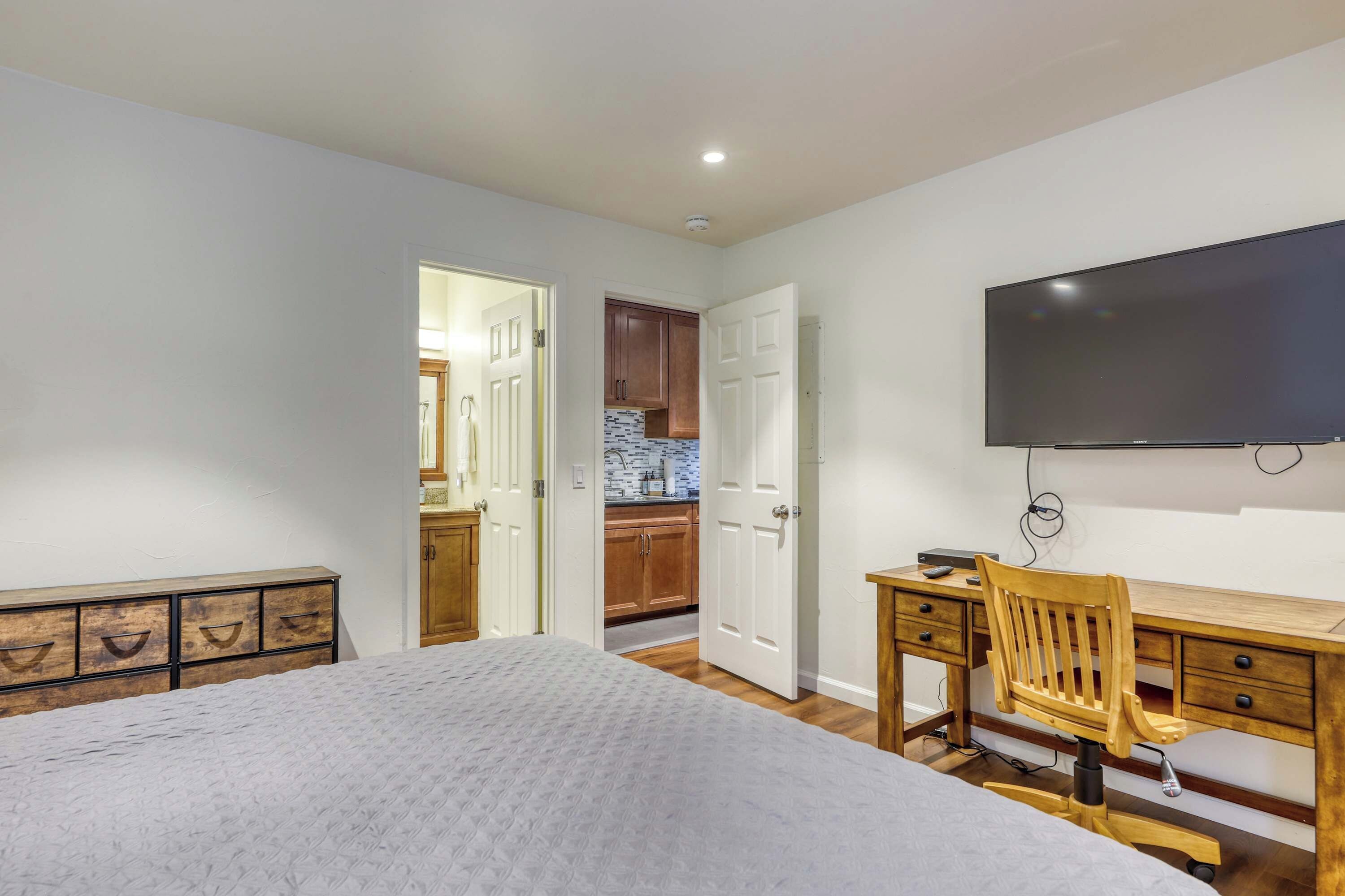 A bedroom with a bed, a desk with a chair, a TV mounted on the wall, and a kitchen visible through an open door.