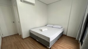 A single bed with a grey mattress, white pillows, and a rolled towel on top. There is a white door on the left and a window on the right.