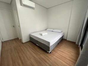 Una cama individual con colchón gris, almohadas blancas y una toalla enrollada encima. Hay una puerta blanca a la izquierda y una ventana a la derecha.